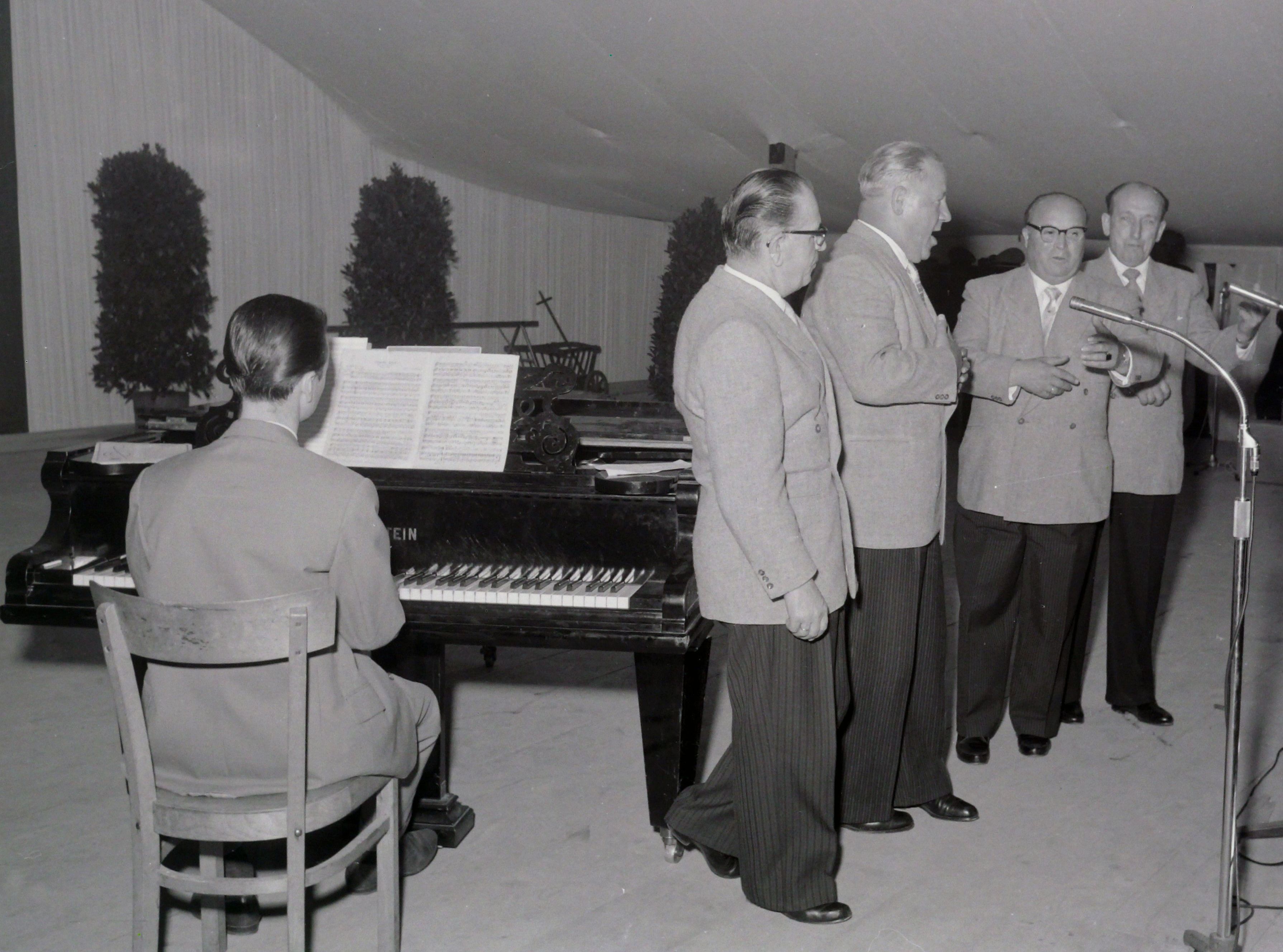 vier Männer singen auf der Bühne, sie werden begleitet von einem Mann am Klavier
